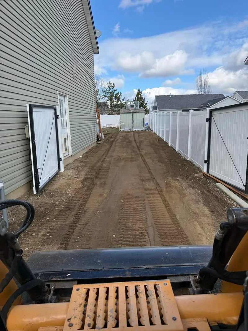 View from a skid steer cab over a freshly graded side-yard driveway along a fence