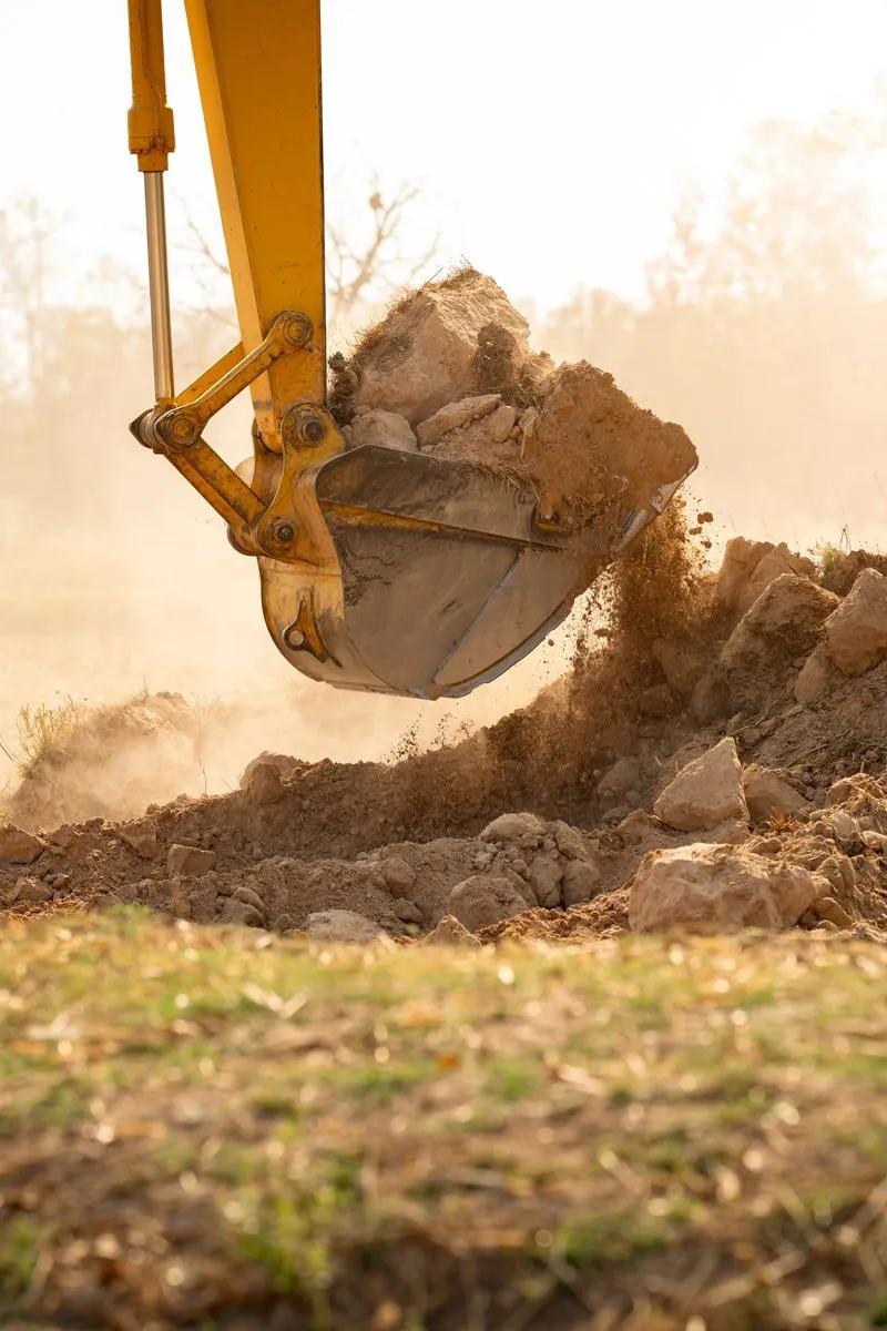 Backhoe bucket digging into dry soil, kicking up dust